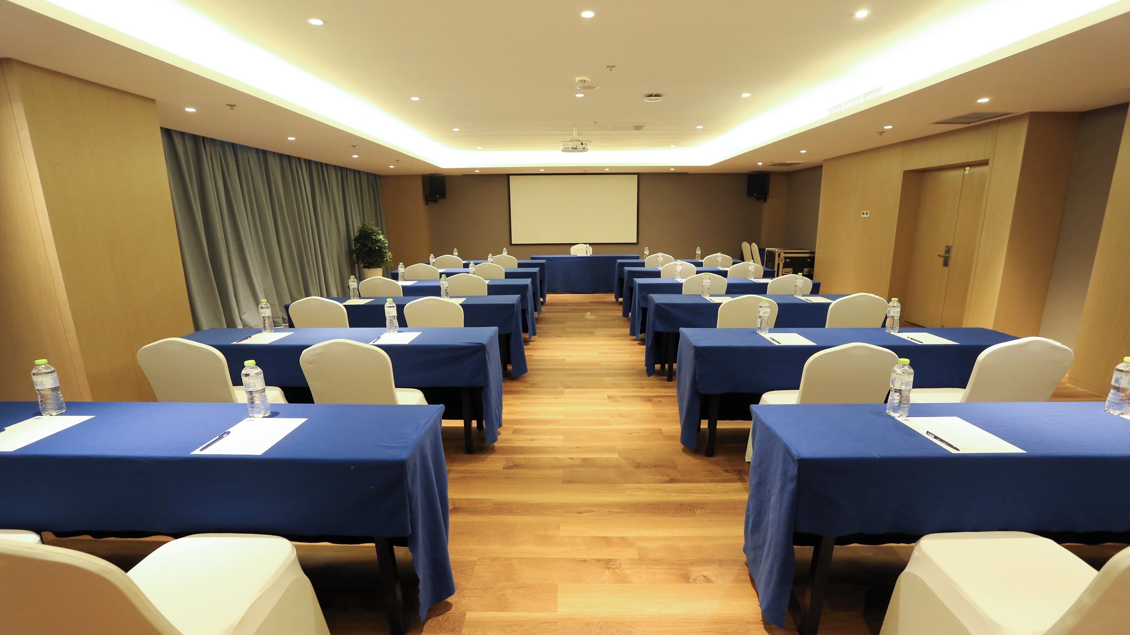Un salón de conferencias con filas de mesas, sillas y una pantalla para proyector.