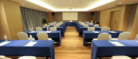 Un salón de conferencias con filas de mesas, sillas y una pantalla para proyector.