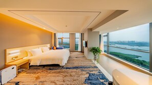 A modern hotel room with a large bed, a desk, and a view of a river and cityscape.