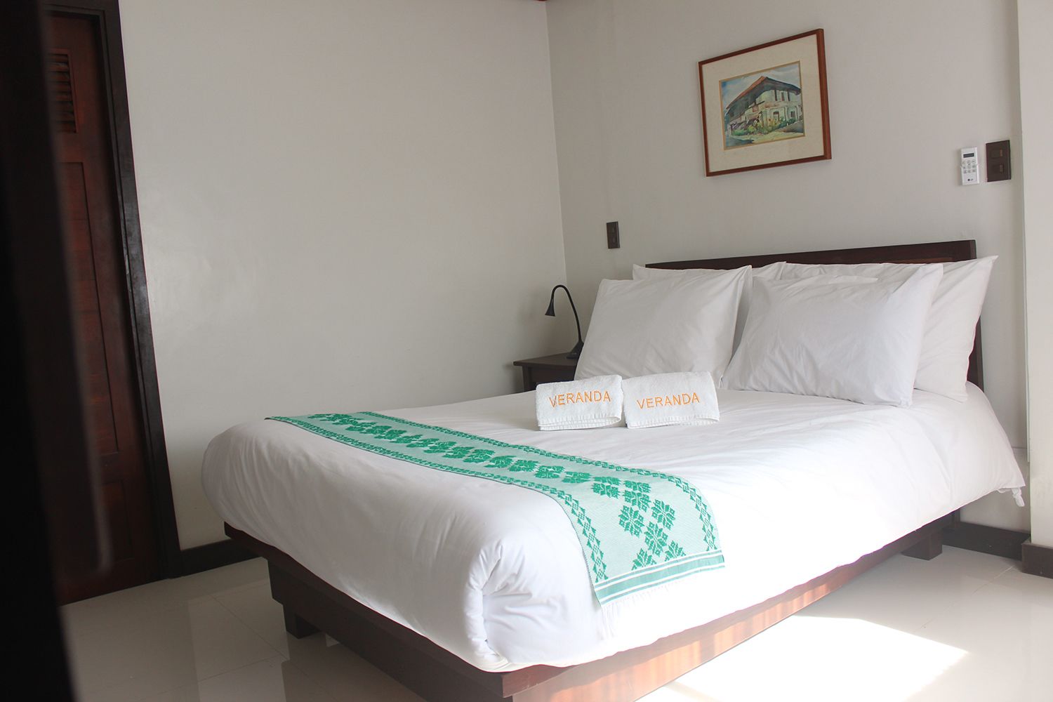 A neatly made bed with white linens and a green patterned throw, two white towels with "VERANDA" on them, a painting on the wall, and a bedside table with a lamp.