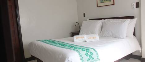 Un lit bien fait, avec du linge de lit blanc et un plaid à motifs verts, deux serviettes blanches ornées de l’inscription « VERANDA », un tableau au mur et une table de chevet avec une lampe.