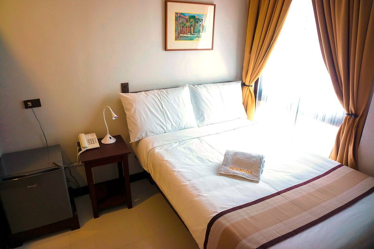 Ett hotellrum med en säng, ett nattduksbord med en telefon och en lampa, ett litet skrivbord och ett fönster med gardiner.