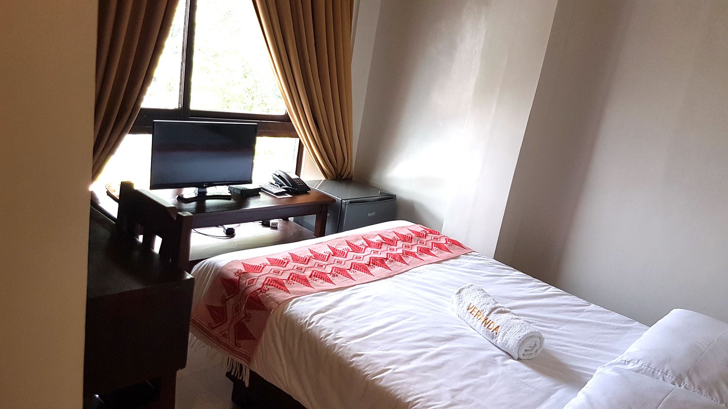A hotel room with a bed, a desk with a computer, a telephone, and a window with curtains.