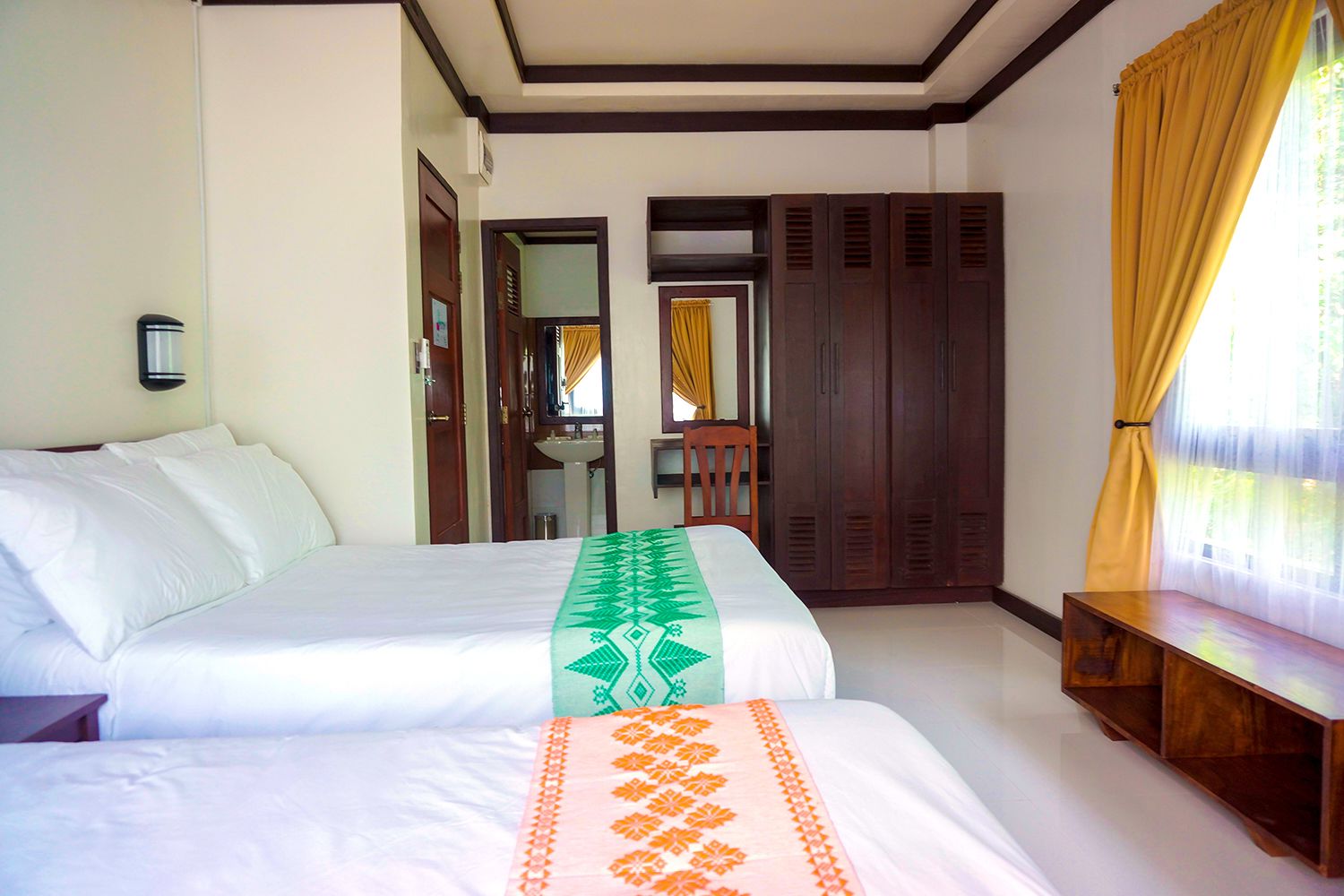 A hotel room with two beds, a wooden wardrobe, a bench, and a bathroom visible through an open door.