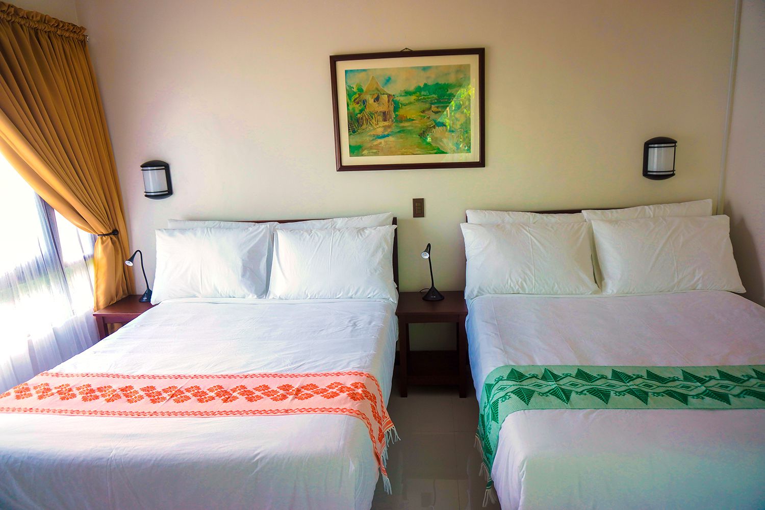 Une chambre d’hôtel avec deux lits, chacun agrémenté de draps blancs et de coussins décoratifs. Un tableau est accroché au mur au-dessus des lits, et deux lampes de chevet sont placées de chaque côté.