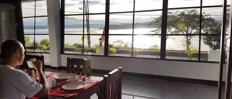 A dining area with a table set for two, large windows with a view of a body of water and greenery, and a person sitting at the table.