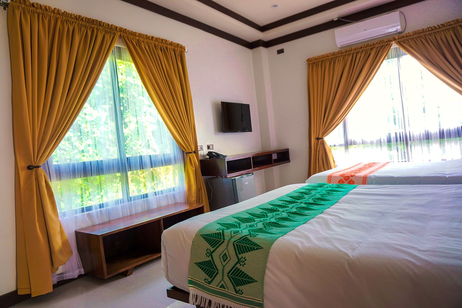 A hotel room with two beds, a desk, a TV, and large windows with curtains.