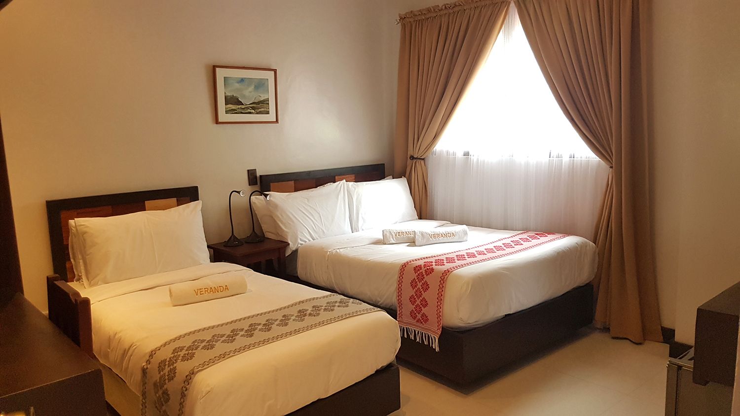 A hotel room with two beds, a nightstand, a lamp, a framed picture on the wall, and a window with curtains.