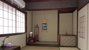 Ein traditioneller japanischer Raum mit Tatami-Boden, einem niedrigen Tisch, Kissen, einem Klappstuhl, einem kleinen Schrank und einer Wandablage mit Bildern.