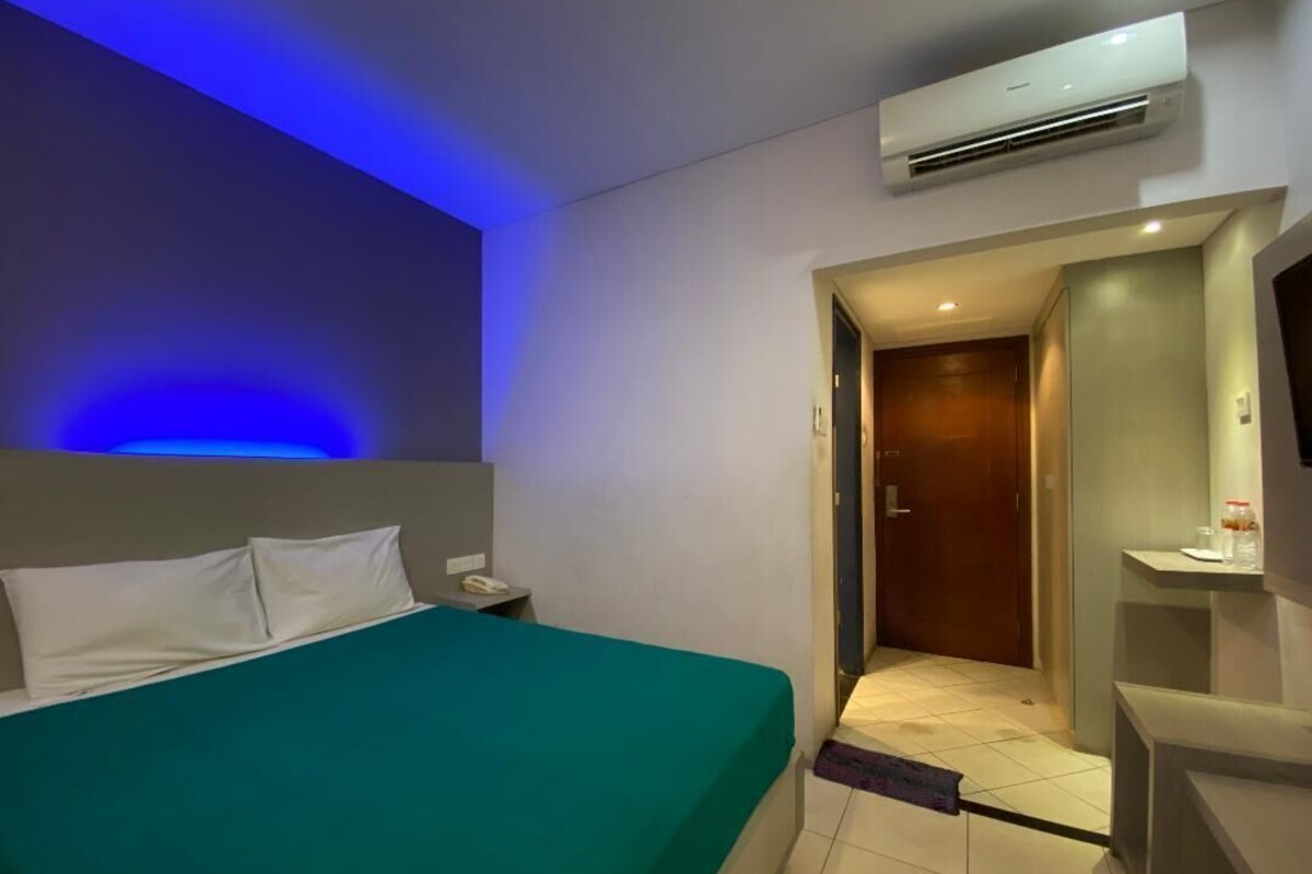 Habitación de hotel con cama, mesita de noche, equipo de aire acondicionado, baño con espejo y puerta que da a otra habitación.