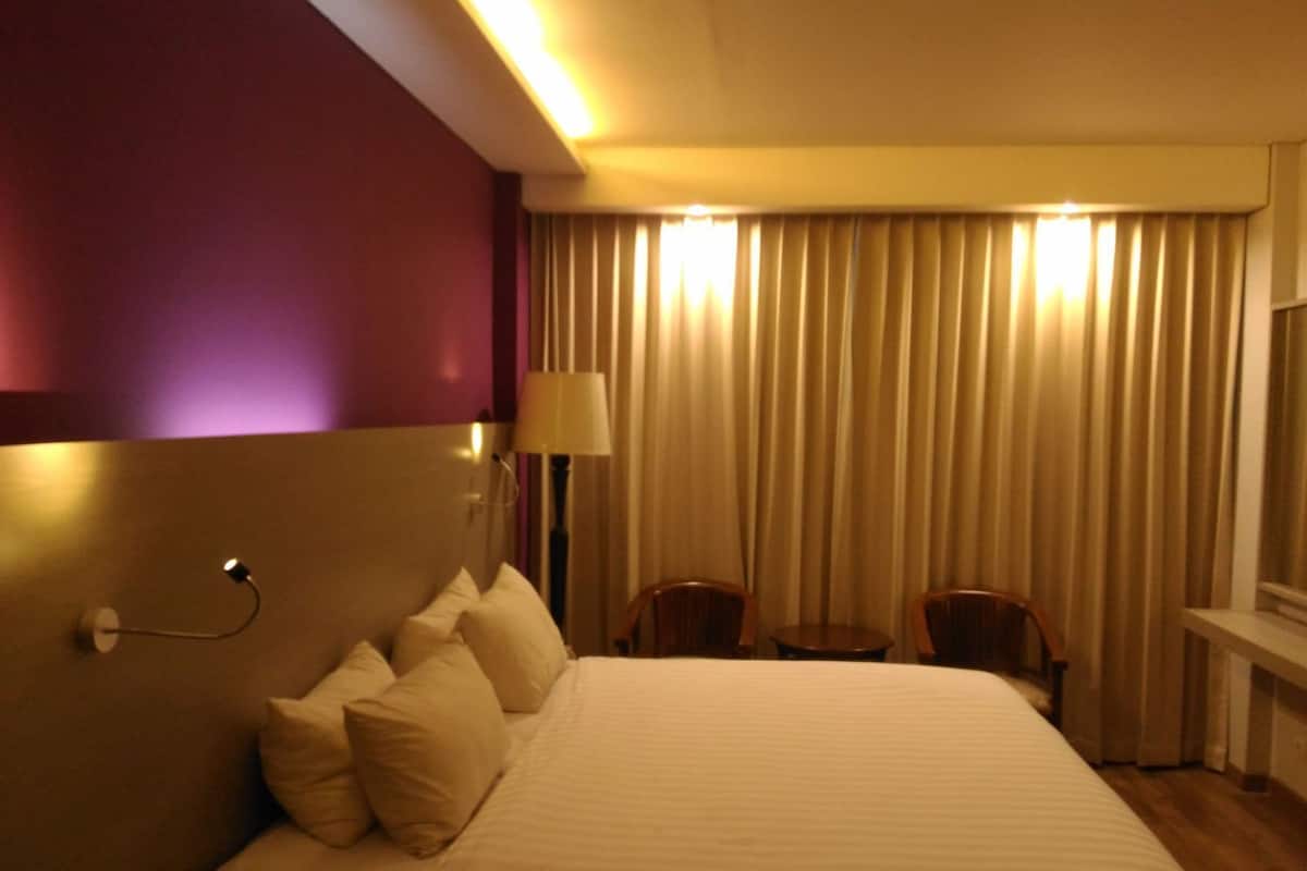 A hotel room with a bed, a desk, two chairs, a lamp, and a window with curtains.