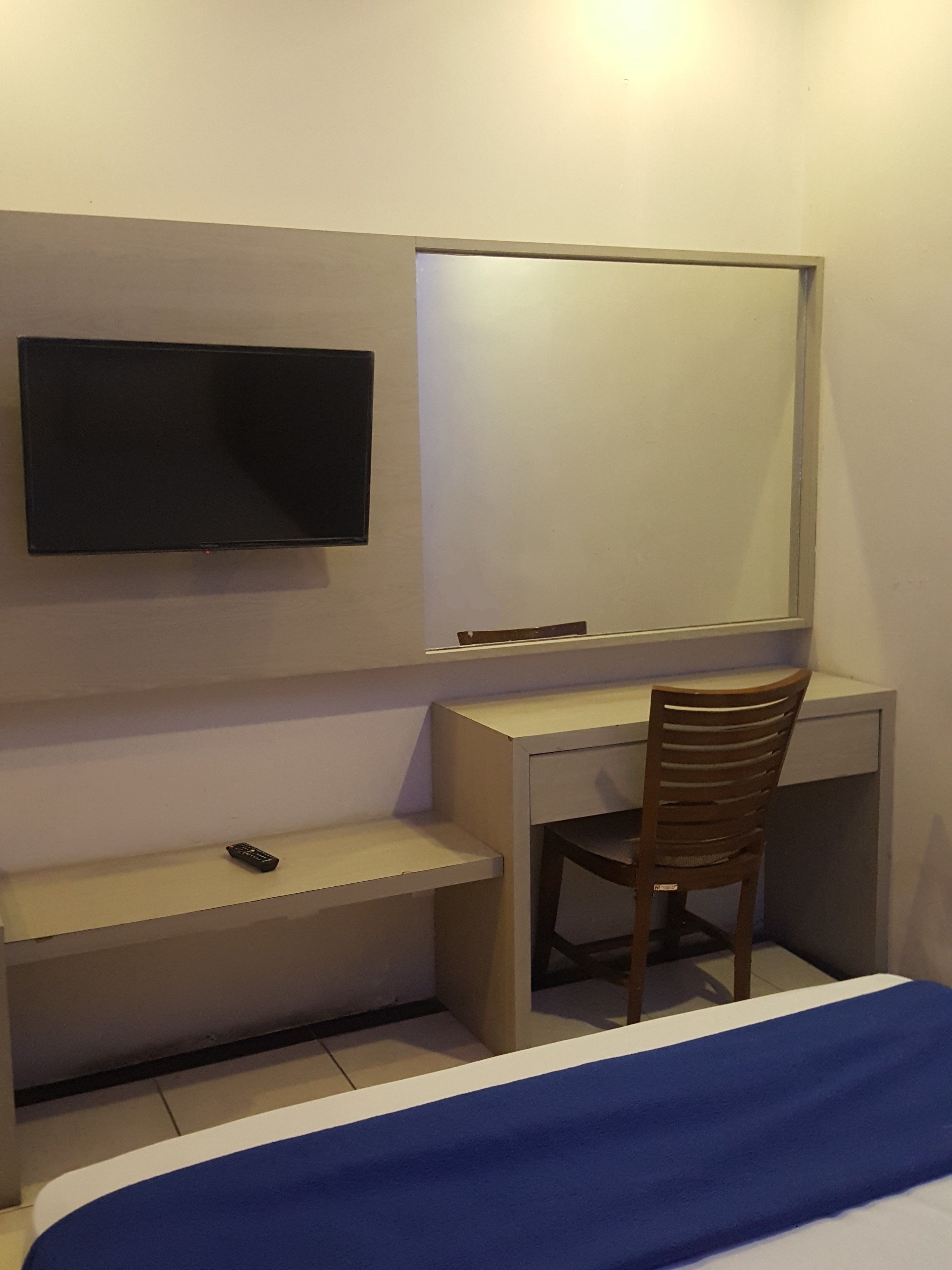 Habitación de hotel con cama, escritorio, silla, televisor montado en la pared y una pequeña estantería.