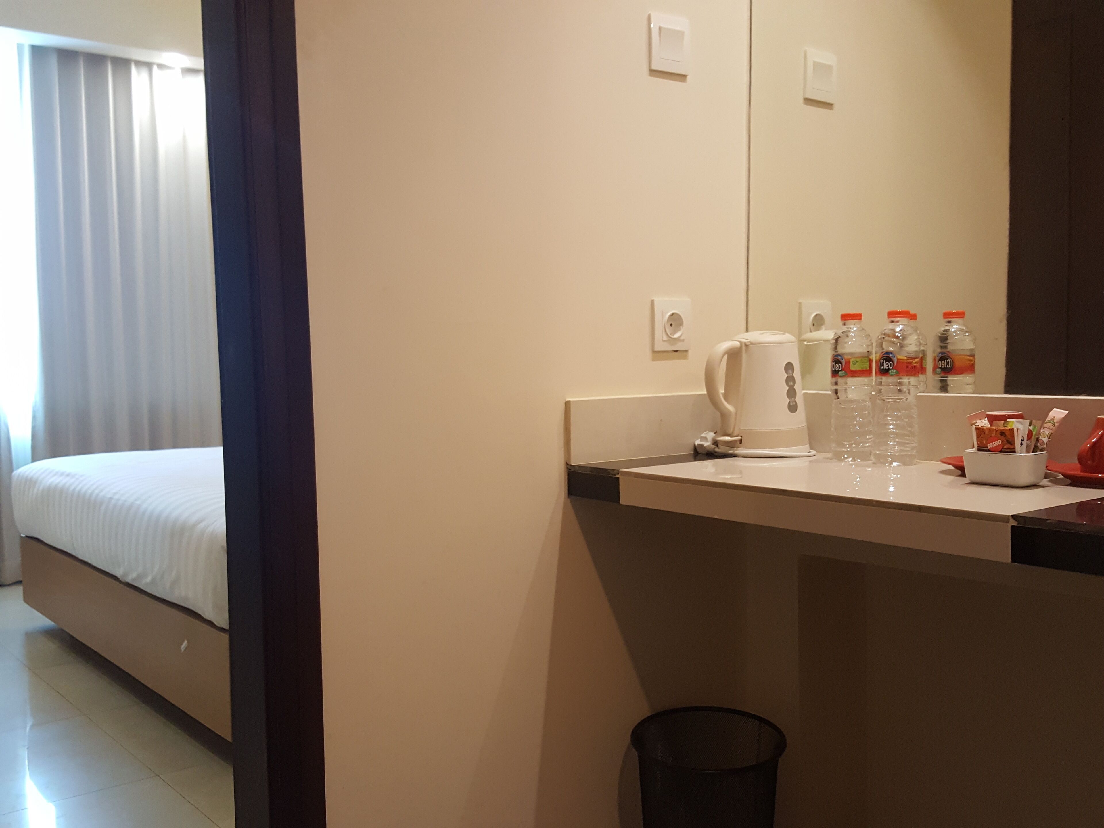 Habitación de hotel con cama, escritorio con cafetera, botellas de agua y un cubo de basura.