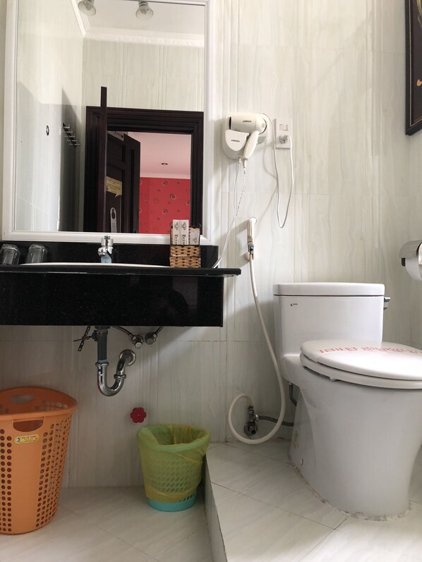 A bathroom with a white toilet, a black sink with a mirror above it, a hairdryer on the wall, and a trash bin. - Linh Phuong 2 Hotel (Can Tho)
