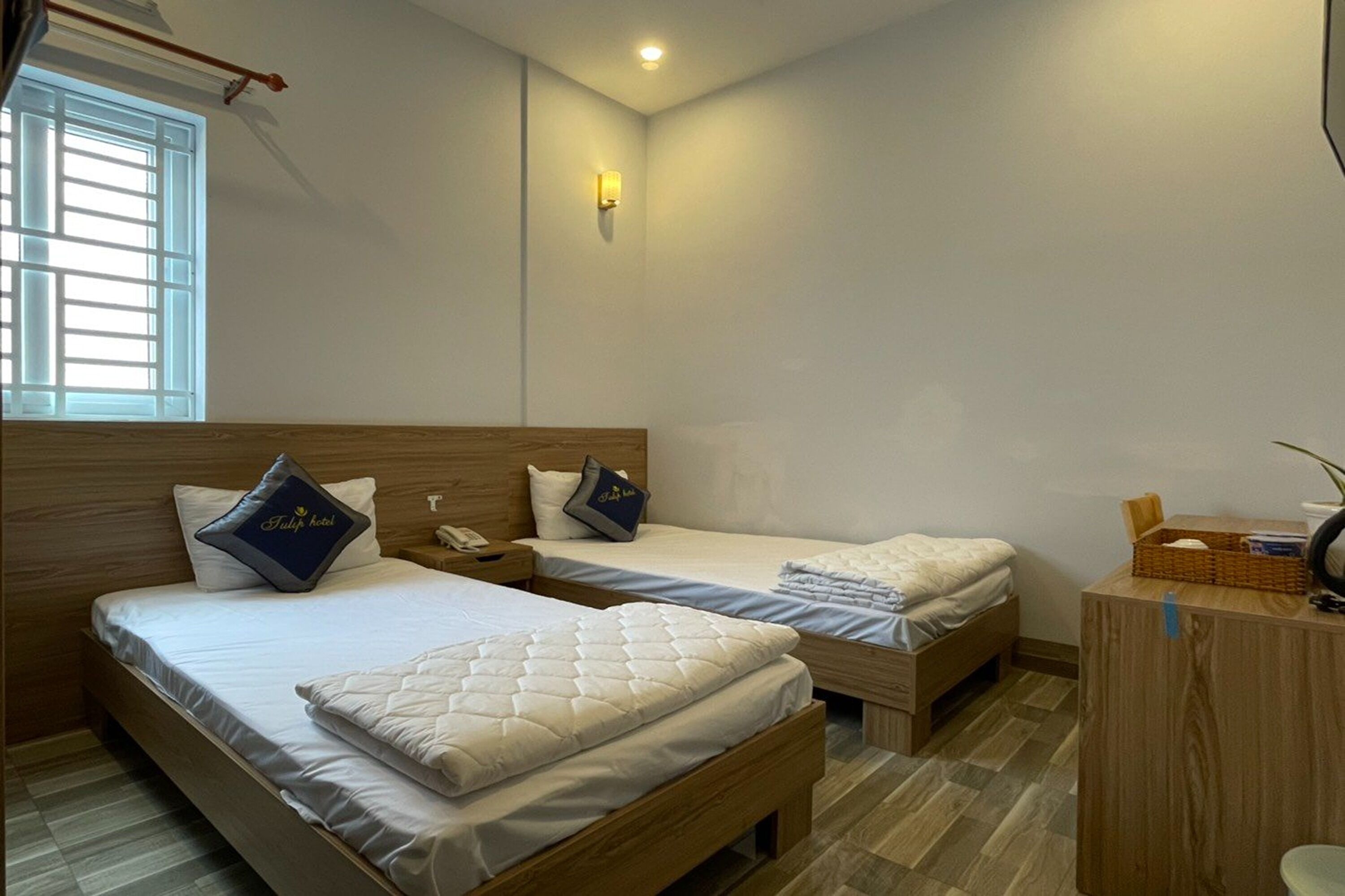 Habitación de hotel con dos camas, cabecero de madera, mesita de noche con lámpara, ventana con persianas y un escritorio pequeño con una cesta y una taza.
