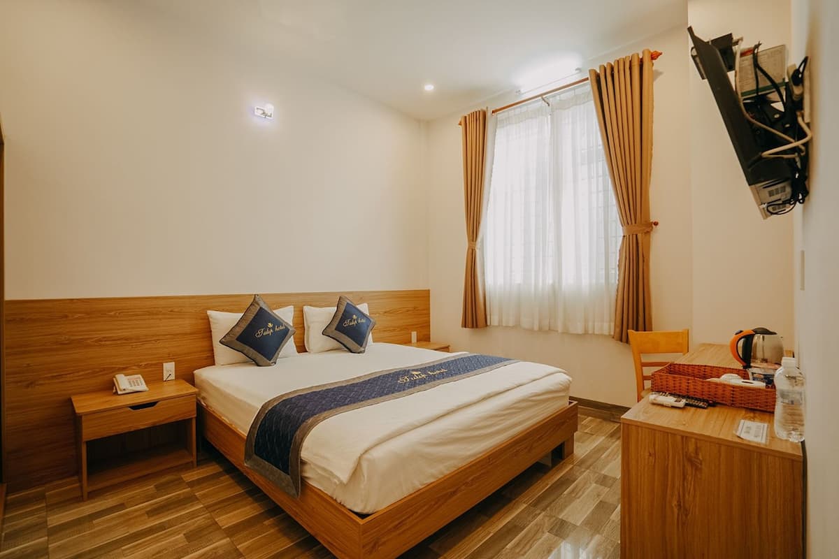 A hotel room with a wooden bed, a nightstand, a desk with a chair, and a window with curtains.