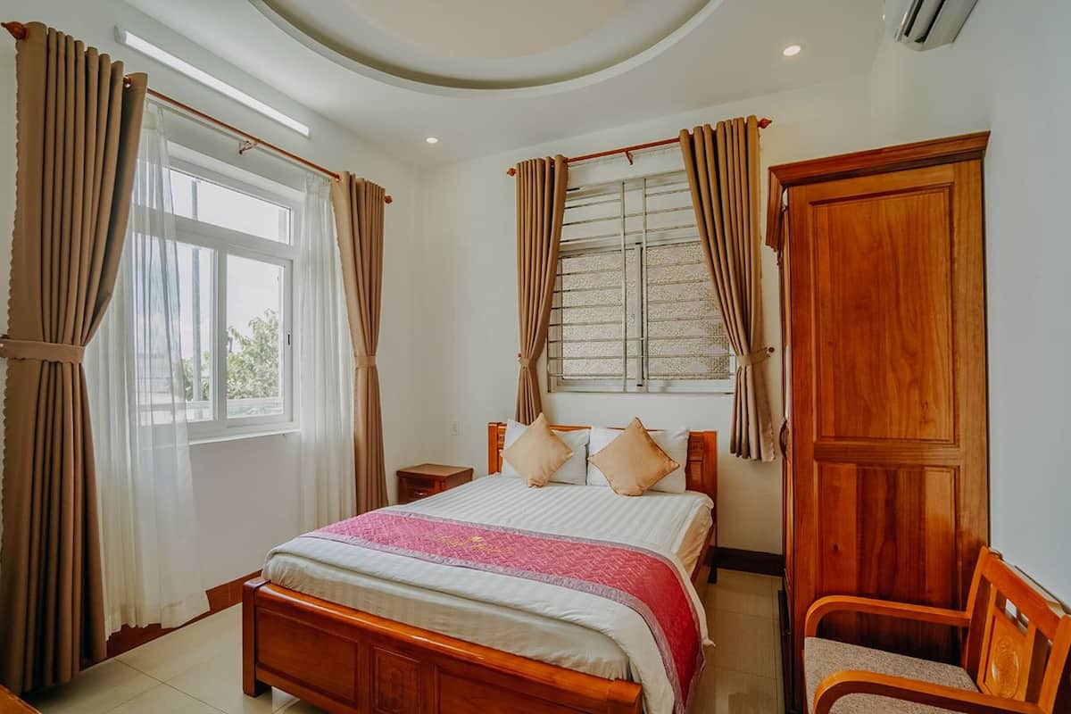A bedroom with a wooden bed, beige curtains, a window with a view, and a wooden wardrobe.