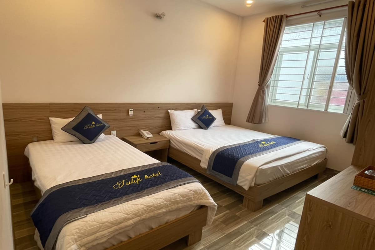 A hotel room with two beds, each with a pillow and a blanket featuring the hotel's logo. There is a bedside table with a phone and a small lamp. A window with curtains is visible, and the room has a wooden headboard and a wooden desk with a basket on it.