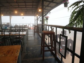 Un restaurante al aire libre con mesas y sillas de madera, barandilla de metal y macetas colgantes de flores.