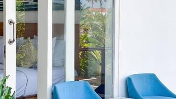 A balcony with two blue chairs and a small table, a glass door leading to a room, and a view of greenery outside.