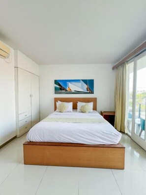 A hotel room with a large bed, a painting on the wall, and a view through the window. - Apple Suite Apartment (Keroboken Kelod)