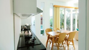 A compact kitchen with a gas stove, kettle, and sink. Adjacent is a dining area with wicker chairs and a table, overlooking a pool. - Apple Suite Apartment (Keroboken Kelod)