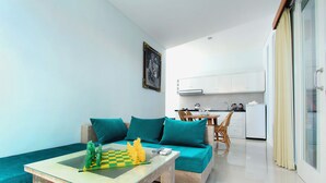 A modern living room with a sofa, a glass-top coffee table, and a chessboard. There is a kitchen area with white cabinets and a dining table with chairs. - Apple Suite Apartment (Keroboken Kelod)