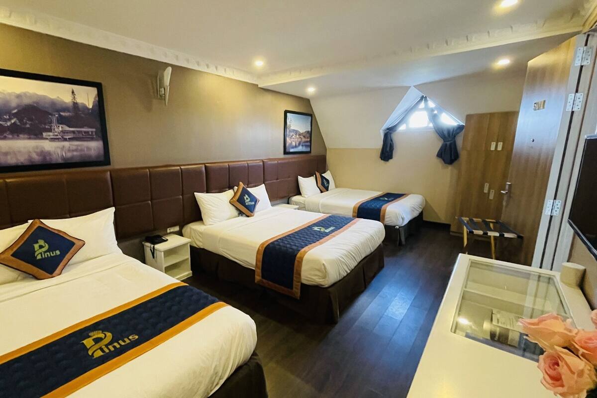 A hotel room with three beds, each with a headboard and a bedspread featuring a logo. There is a framed picture on the wall, a window with curtains, and a wooden door.