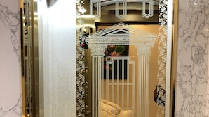 Un ascensore lussuoso con pareti in marmo e pavimento decorato, che conduce a una stanza con un letto e uno specchio ornamentale.