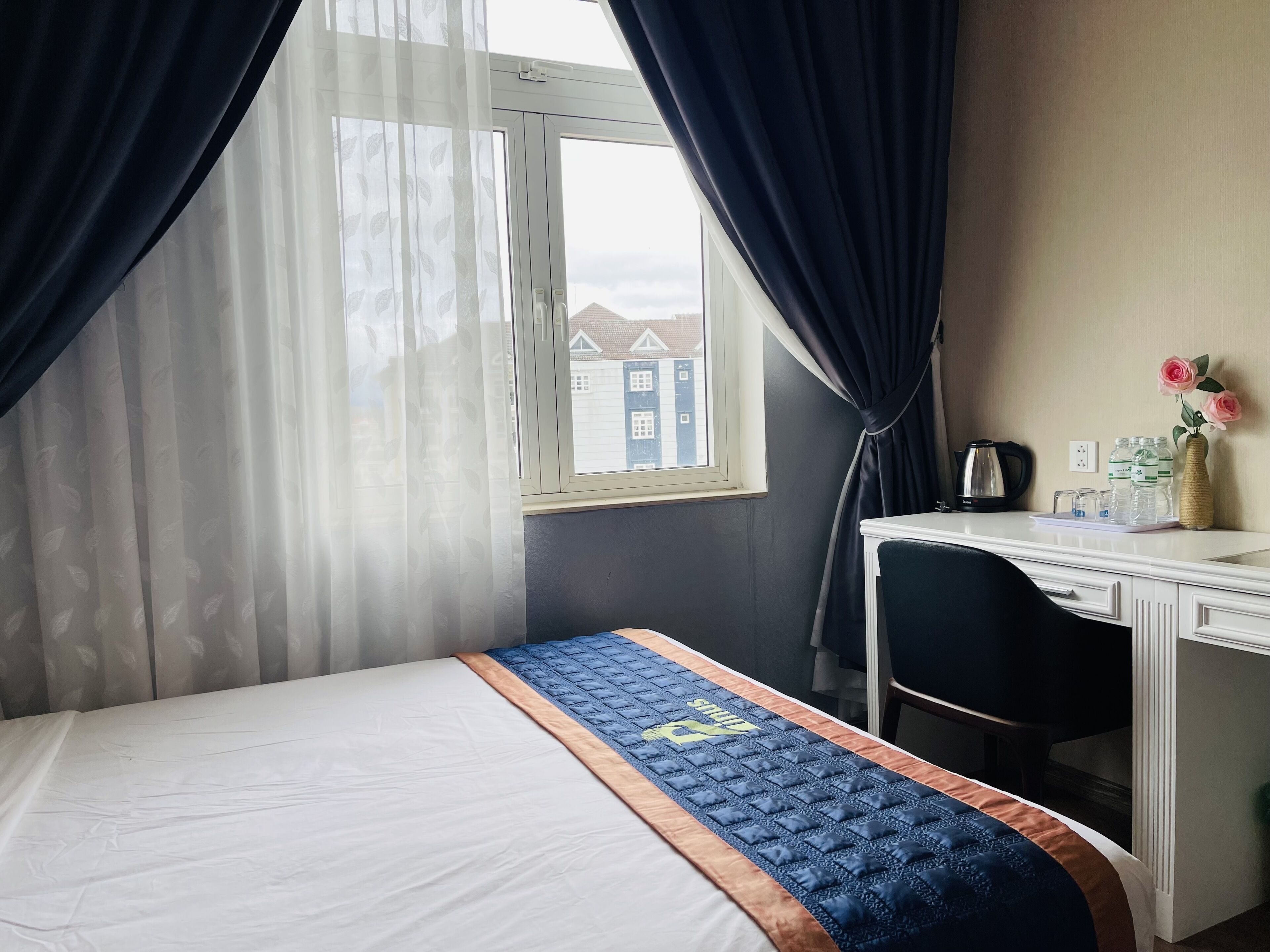 Una habitación de hotel con una cama, un escritorio, una silla, una ventana con cortinas y un jarrón con flores.