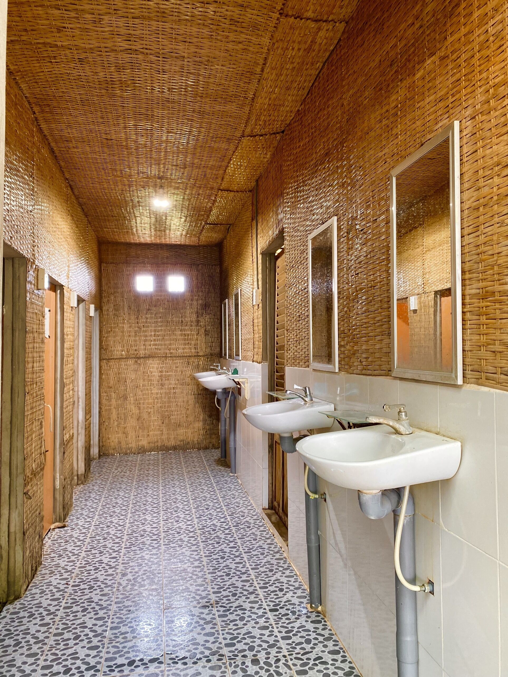 A narrow hallway with a tiled floor and a ceiling made of woven material. There are three sinks along the right wall, each with a mirror above.