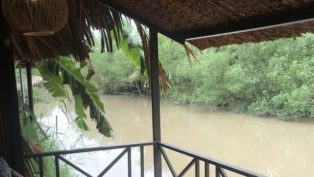 Área externa coberta com cadeiras de bambu e guarda-corpo com vista para um rio.