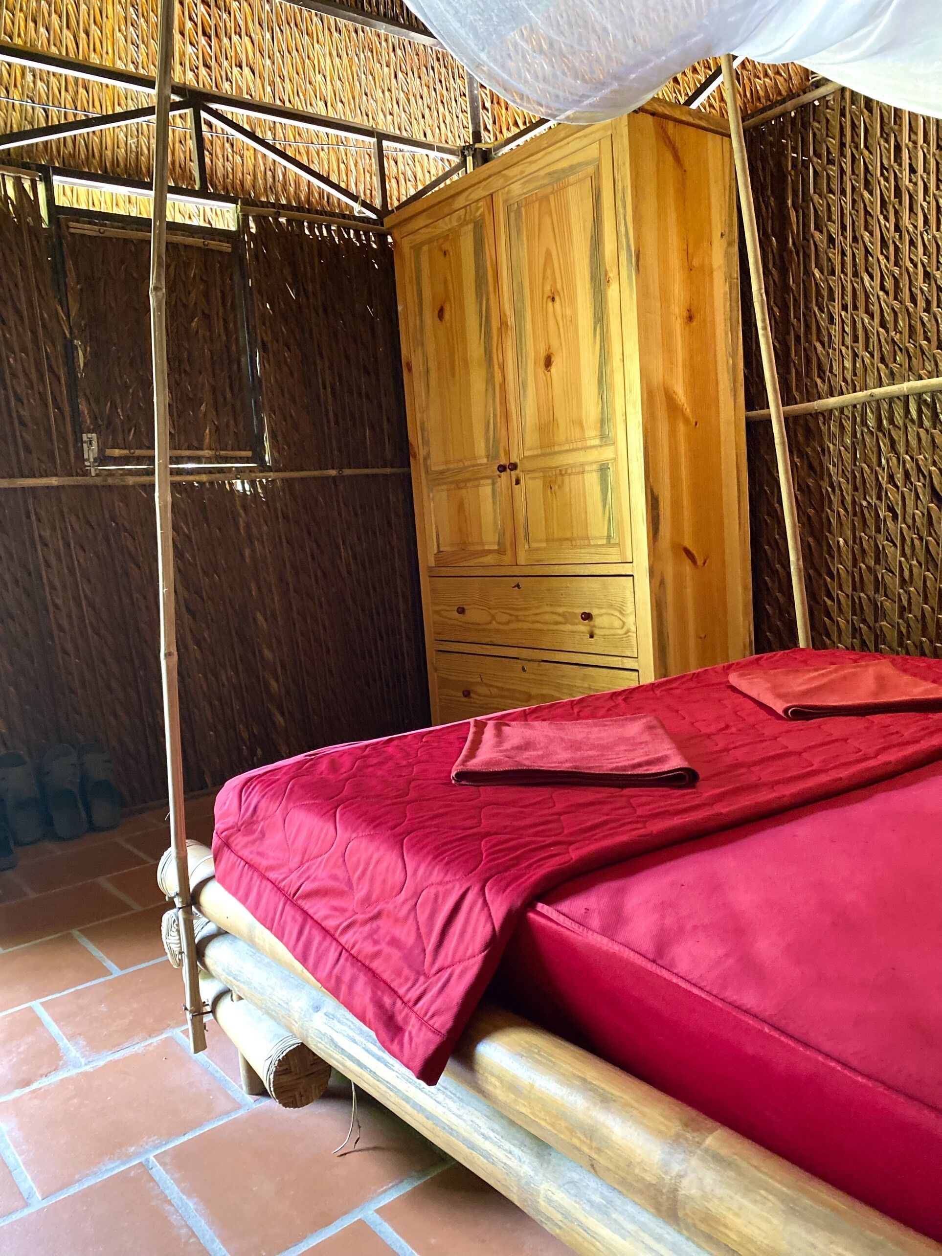 A room with a wooden wardrobe, a bed with a red bedspread, and a thatched roof structure.
