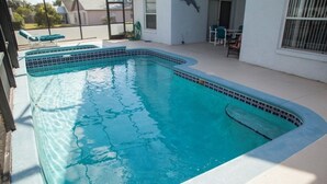 Et rektangulært pool med flisekant, omgivet af en overdækket patio med et bord og stole.