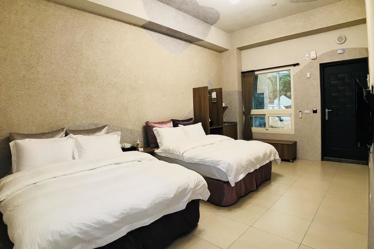 Habitació d'hotel amb dos llits, una finestra amb cortines, una porta i una taula petita amb una làmpada.