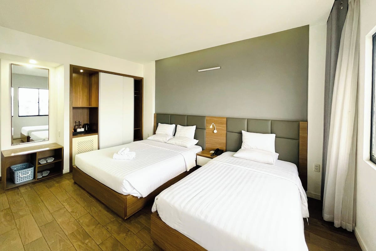 A hotel room with two beds, a large mirror, a wardrobe, a bedside table with a lamp, and a window with curtains.