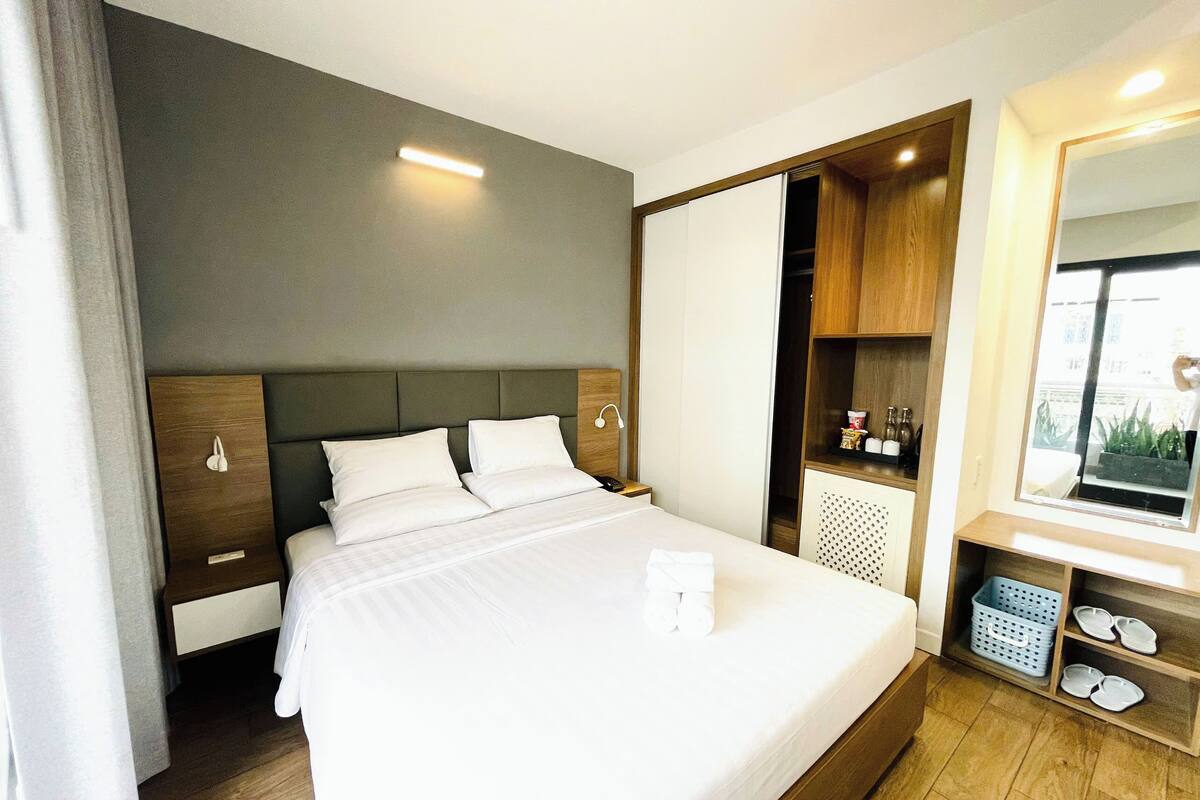 A modern hotel room with a large bed, a wooden headboard, a built-in wardrobe, and a small seating area with a mirror and shelves.