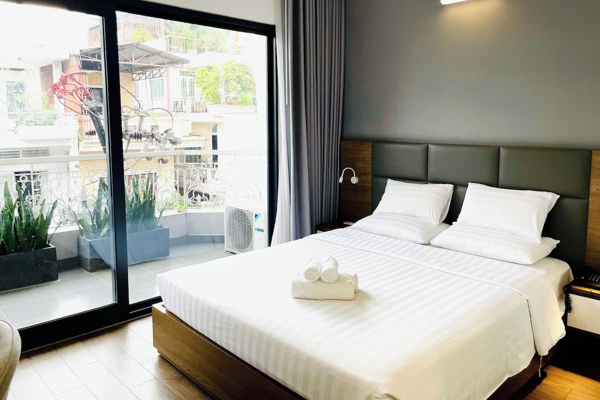 A hotel room with a large bed, a nightstand, a modern headboard, a window with a view of the outside, and a potted plant.