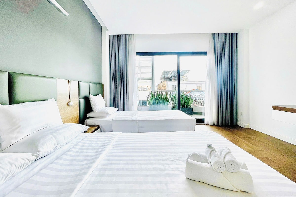 A hotel room with two beds, a large window with curtains, and a view of buildings outside.