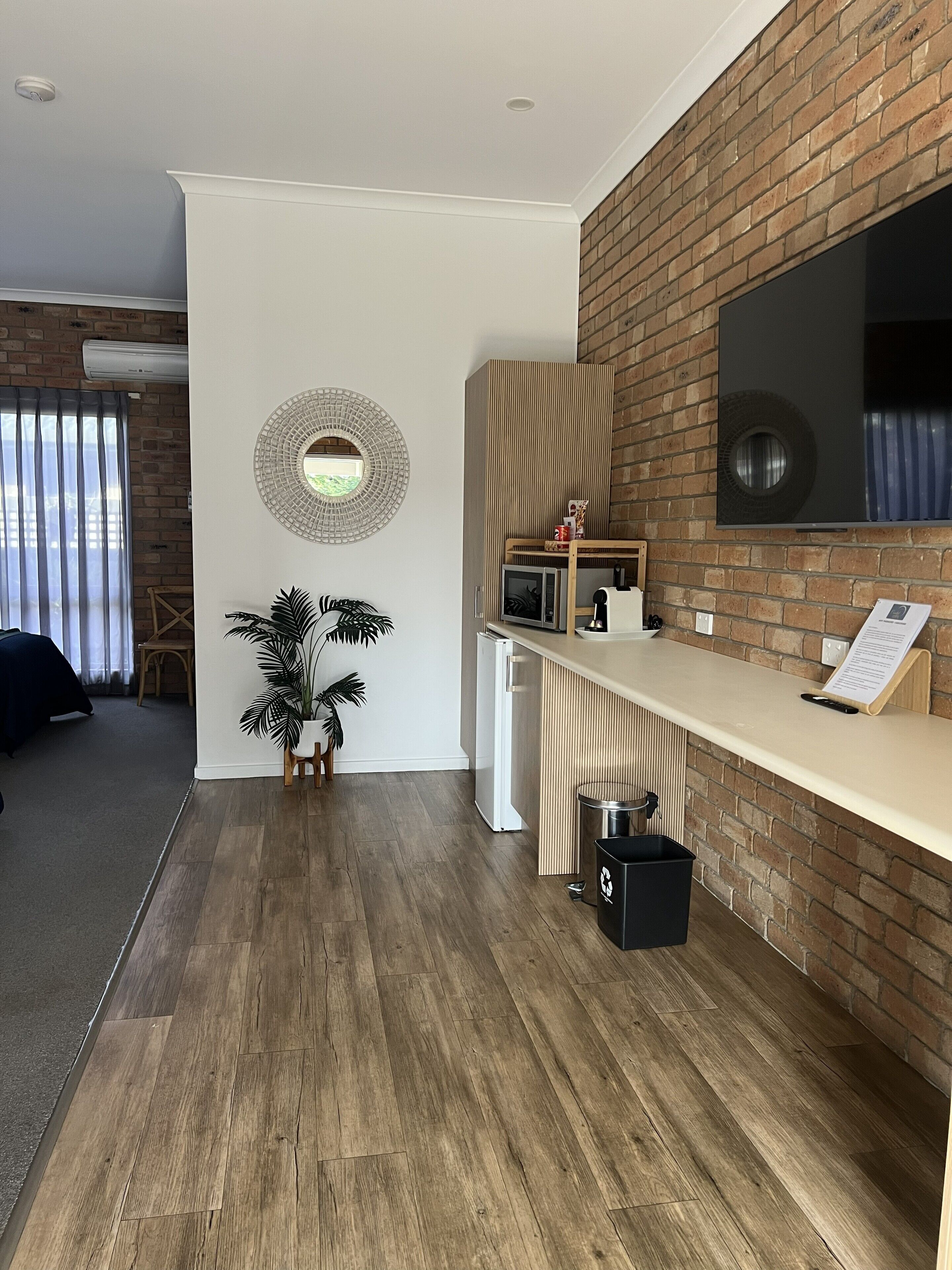 Una sala de estar moderna con pared de ladrillo, un televisor grande, un sofá blanco, una mesita con una planta y piso de madera.