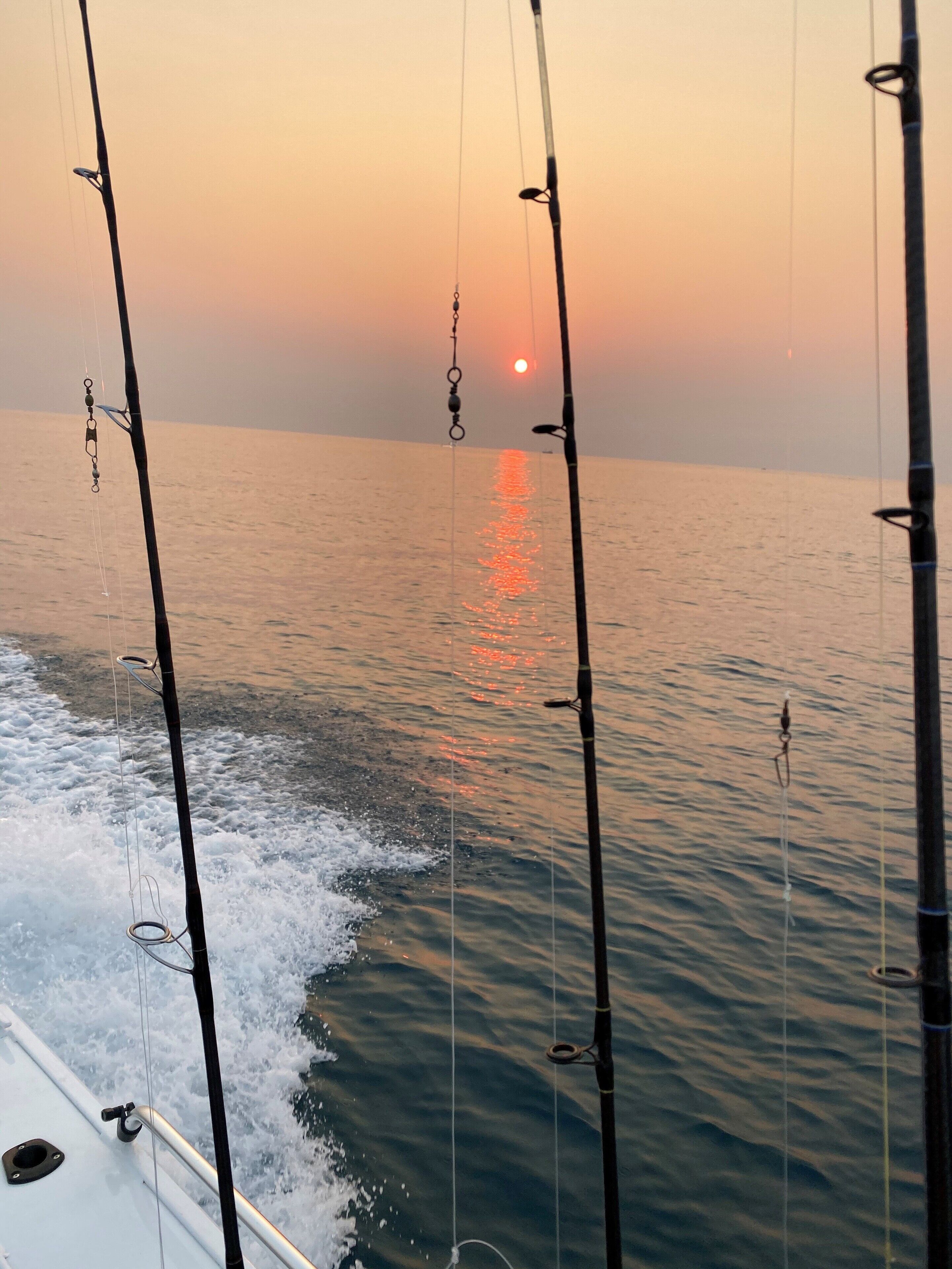 A calm sea with fishing rods extending into the water, a boat's wake visible, and a sunset in the background.