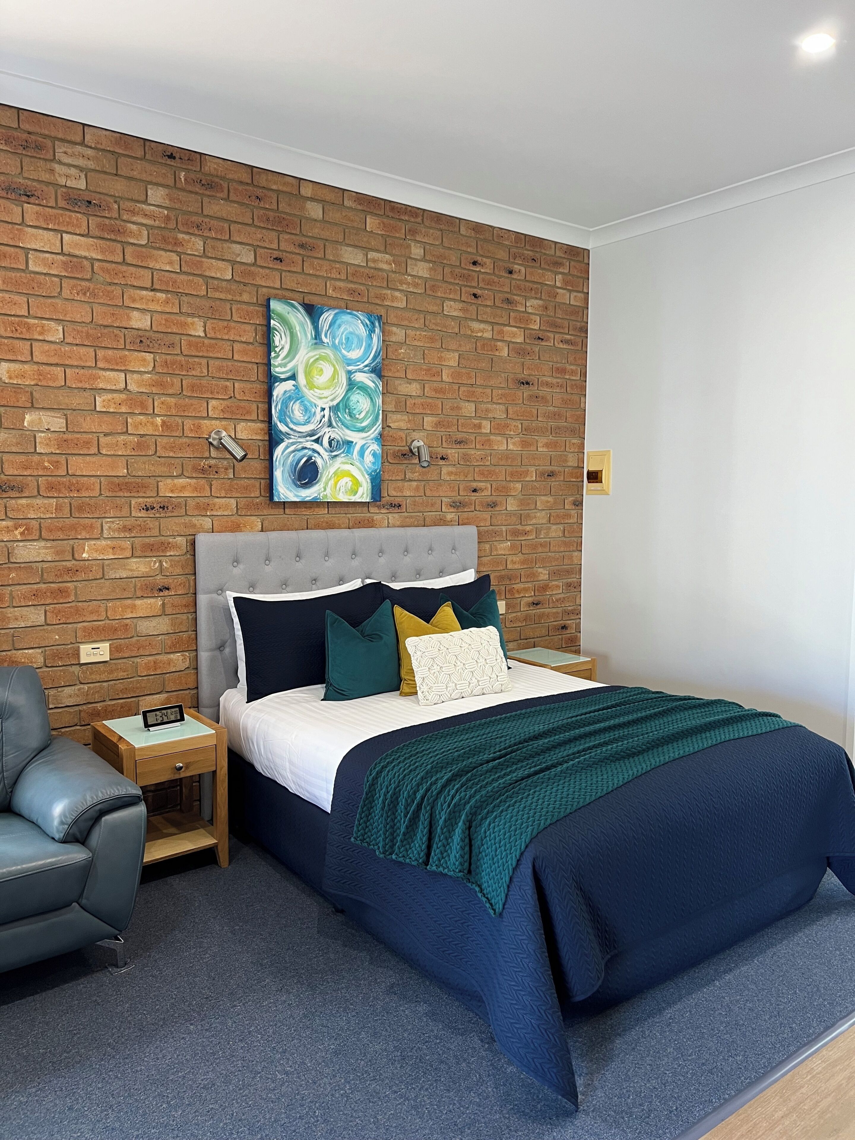 A bedroom with a brick wall, a bed with a grey headboard, a grey armchair, a bedside table with a clock, and a painting above the bed.