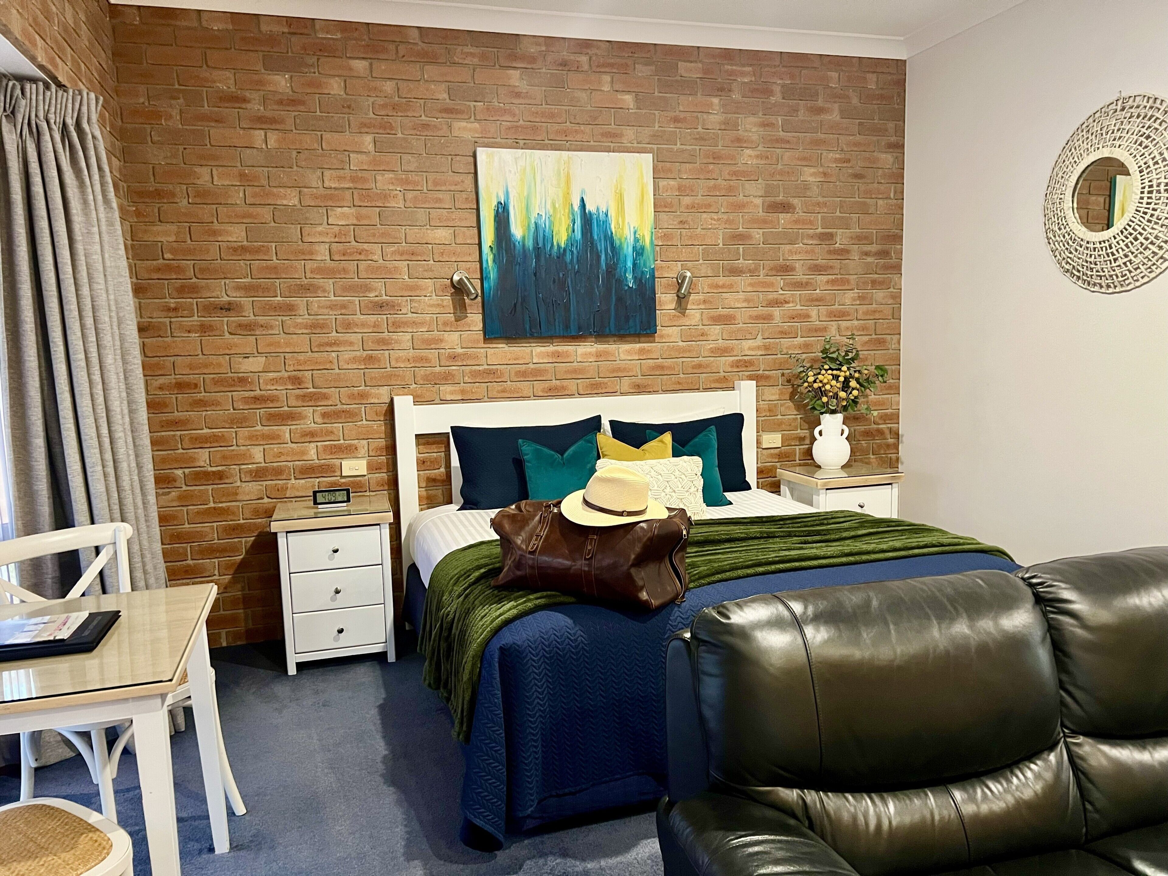 A hotel room with a brick wall, a bed with a white headboard, a nightstand, a desk, a leather chair, and a painting on the wall.