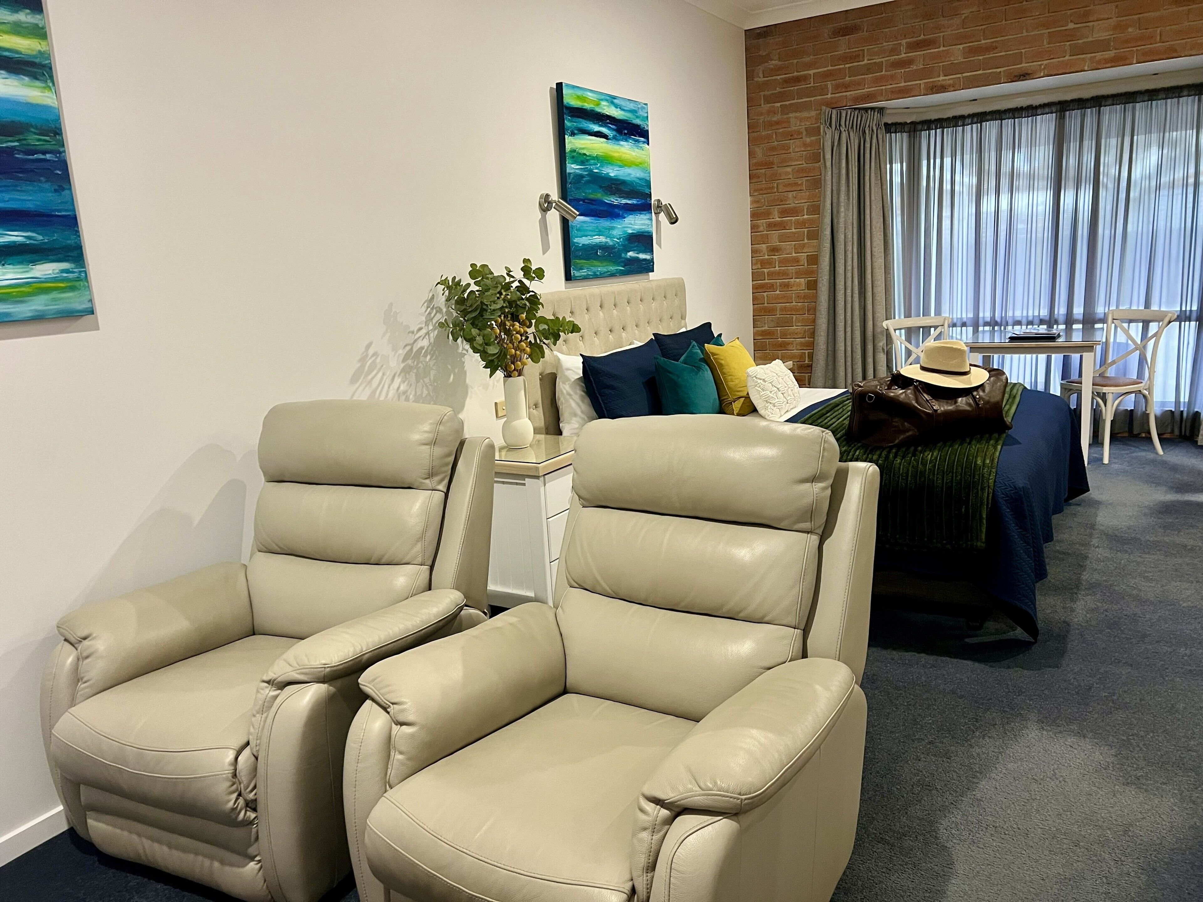 A hotel room with a bed, two leather armchairs, a side table with a plant, a painting, and a view through a window with curtains.