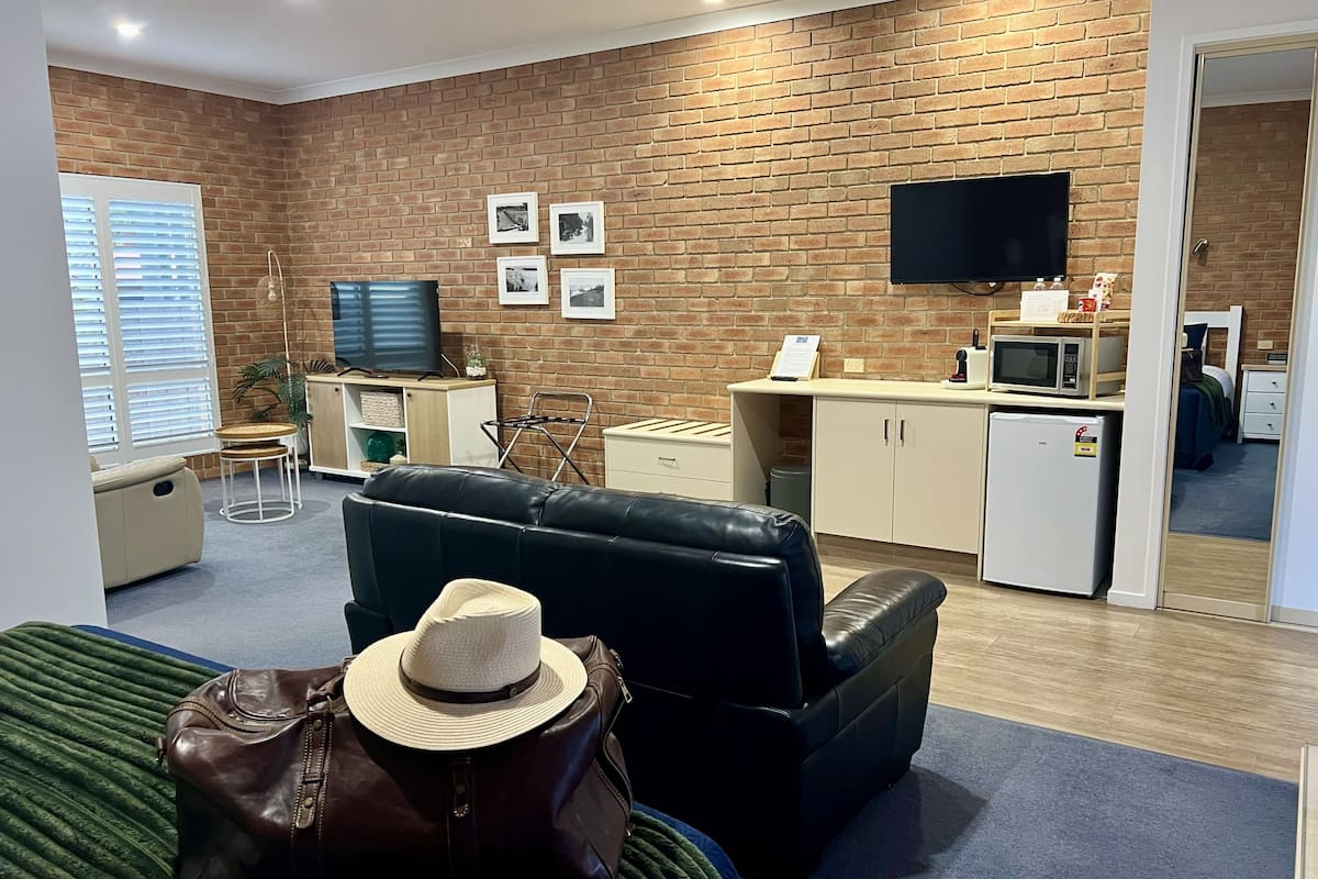 Une chambre d’hôtel avec un mur en briques, un téléviseur à écran plat, une petite kitchenette, un canapé, un lit et une valise.