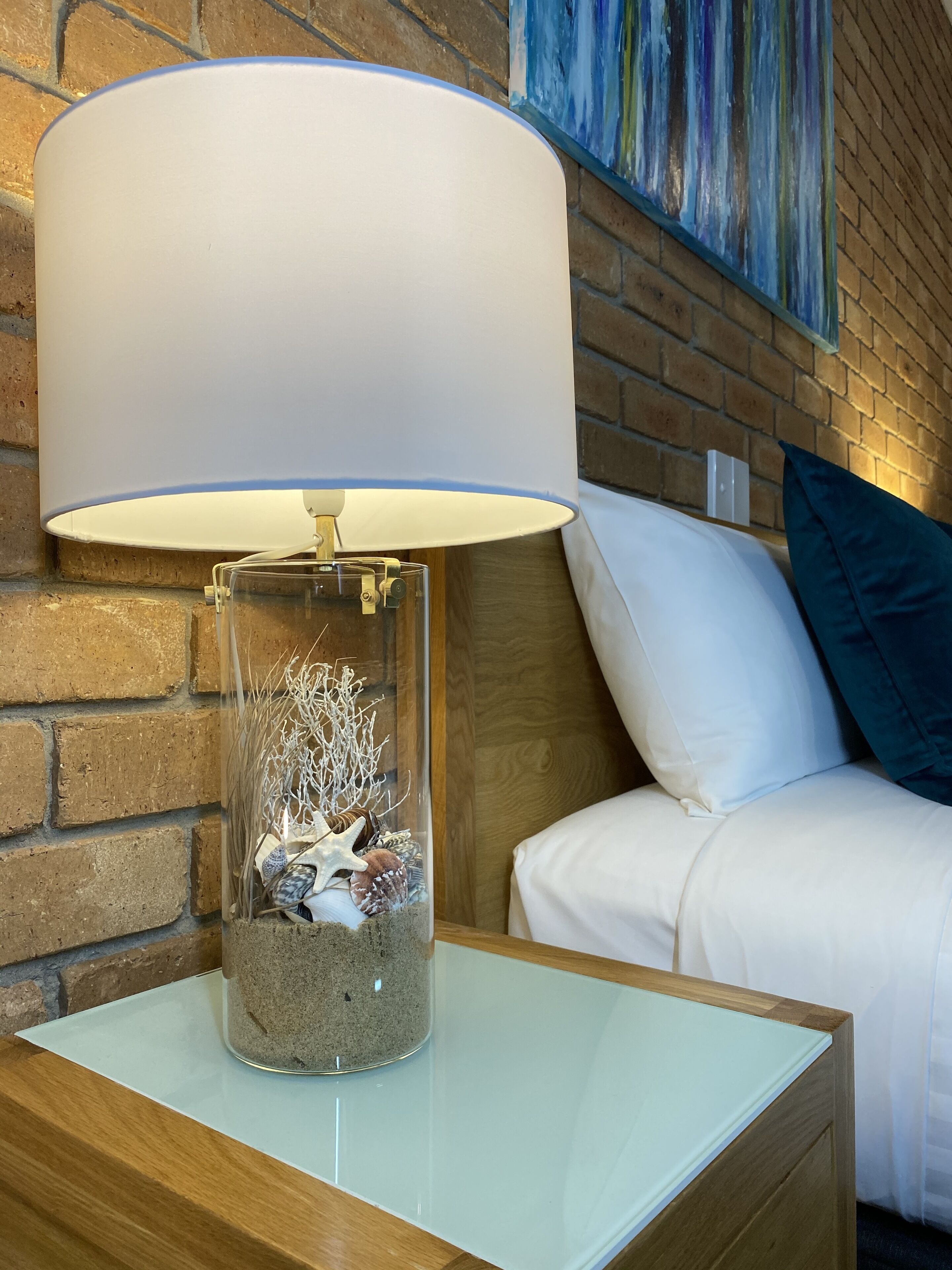 Une lampe en verre transparent avec un abat-jour blanc, posée sur une table de chevet en bois, devant un mur en briques, et une peinture accrochée au-dessus.
