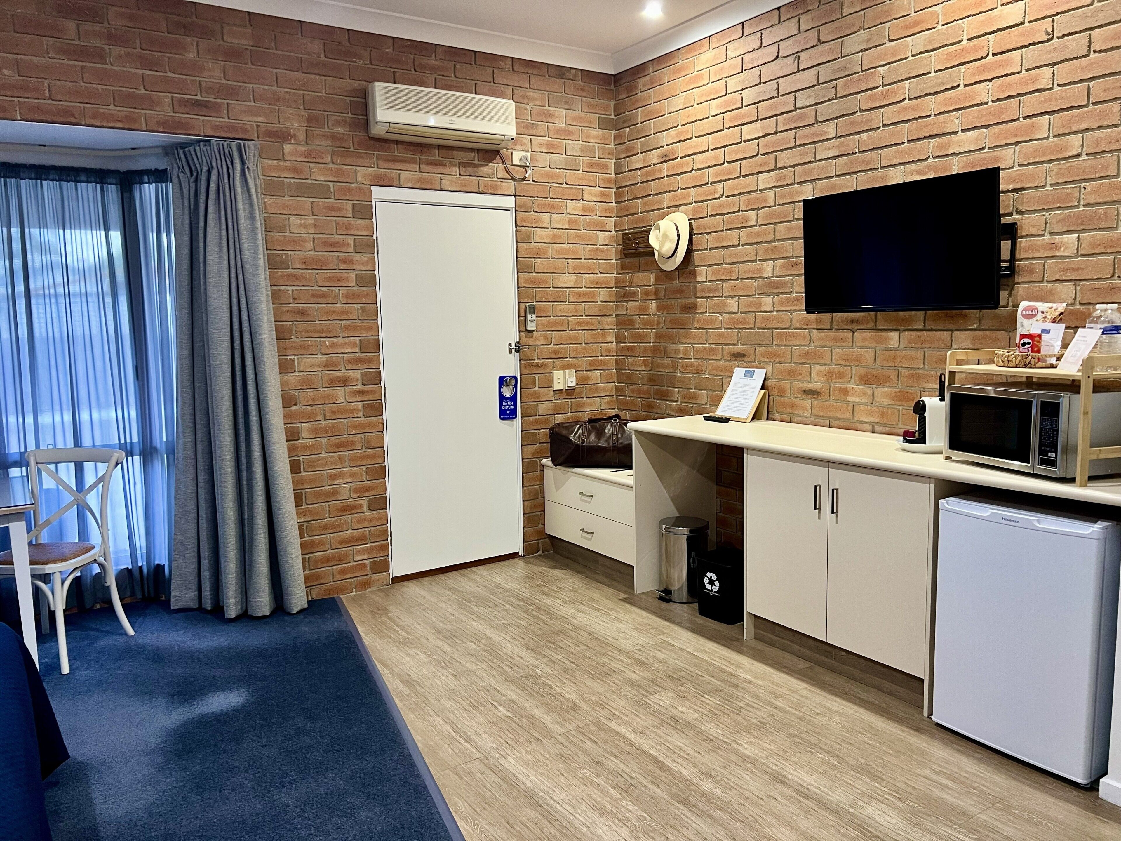 A hotel room with a brick wall, a flat-screen TV, a kitchenette with a microwave, a small refrigerator, a trash bin, and a chair.