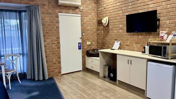 A hotel room with a brick wall, a flat-screen TV, a kitchenette with a microwave, a small refrigerator, a trash bin, and a chair.