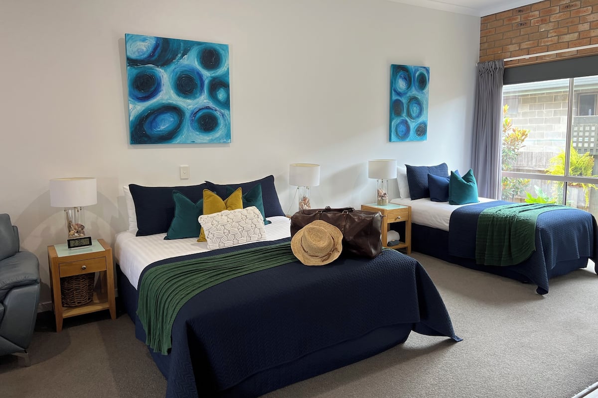 Une chambre d’hôtel avec deux lits, une chaise, une table de chevet, une lampe et des œuvres d’art accrochées aux murs.