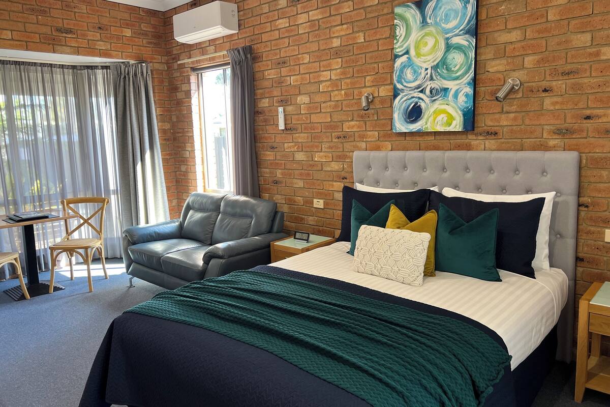 Une chambre d’hôtel avec un mur en briques, une tête de lit rembourrée de couleur grise, un lit avec plusieurs oreillers, un fauteuil en cuir gris, une petite table et une fenêtre avec des rideaux.